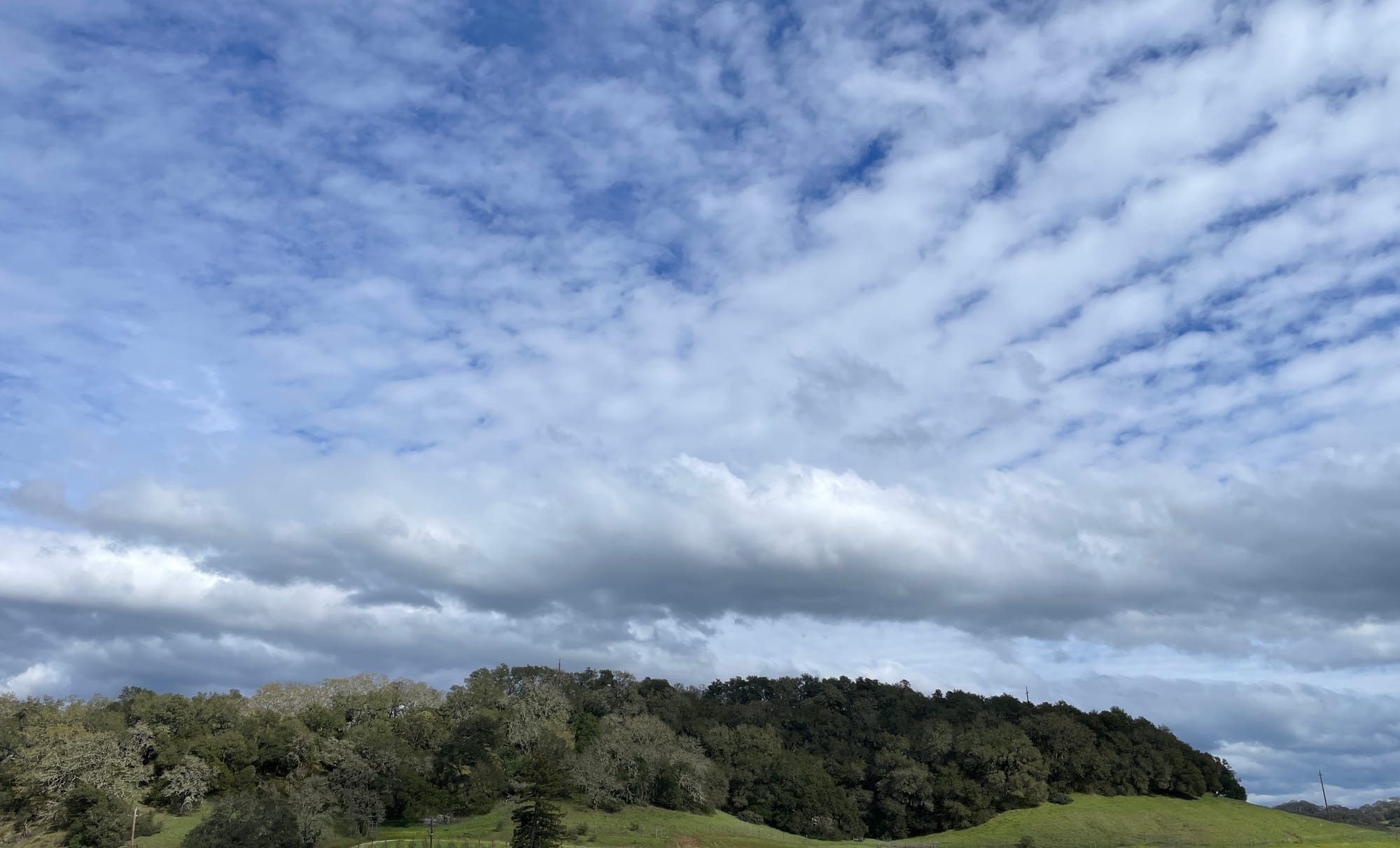 Blue sky peeking through white clouds over a low green hill. 